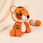 A crocheted tiger plush toy in orange and white with black stripes, posed next to a matching orange yarn ball on a warm beige background.