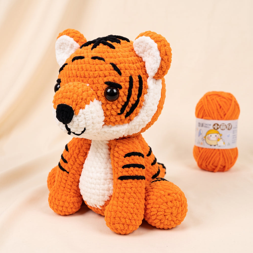 A crocheted tiger plush toy in orange and white with black stripes, posed in front of a small ball of orange yarn on a soft beige background.