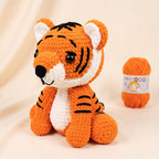 A crocheted tiger plush toy in orange and white with black stripes, posed in front of a small ball of orange yarn on a soft beige background.