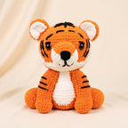 A crocheted tiger amigurumi toy sitting upright, featuring orange yarn with black stripes, a white chest and inner ears, and black embroidered facial details, set against a soft beige draped fabric background.