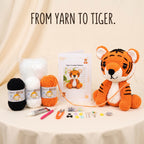 A crocheted tiger toy sits on a draped table beside a booklet labeled 'Tiger Crochet Pattern,' several balls of yarn in black, white, and orange, and small crafting tools. The text 'FROM YARN TO TIGER.