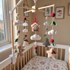 A wooden baby crib with a white headboard and footboard, featuring a hanging crocheted mobile with a gingerbread man, Christmas tree, house, clouds, bells, and red pom-poms. The crib is dressed in a patterned cream sheet, and the room has a warm beige wall with framed art.