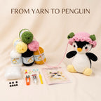 A crocheted penguin toy kit displayed on a beige fabric background with the text 'FROM YARN TO PENGUIN'. The main product is a white and black crocheted penguin wearing a pink and green hat, alongside various yarn balls in green, yellow, pink, black, and white, beads, thread, a small knife, and a pattern booklet.