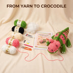 Crocheted yarn kit for making a watermelon crocodile, featuring a finished pink and green crocheted crocodile toy, multiple balls of yarn in green, black, cream, and pink, cotton stuffing, a tube of Stitch brand yarn glue, scissors, and a crochet pattern booklet on a beige fabric background with the text 'FROM YARN TO CROCODILE'.
