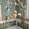 A handmade crocheted baby mobile with a wooden arched frame, featuring a yellow duckling, green frog, blue dragonfly, white lotus, snail, and brown cattails, displayed in a nursery crib.