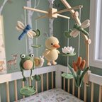 A handmade crocheted baby mobile with a wooden arched frame, featuring a yellow duckling, green frog, blue dragonfly, white lotus, snail, and brown cattails, displayed in a nursery crib.