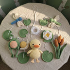 A set of handmade crocheted spring-themed ornaments arranged on a beige fabric-covered stool. The set includes a yellow crocheted duckling, green lily pads, a blue dragonfly, a green dragonfly, a white flower, a brown snail, and orange tulip-like stems, all connected by white cords.