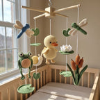 A handmade crocheted baby mobile with a wooden top frame, featuring a yellow duckling, green frog, blue dragonfly, white lotus, and orange reeds, hanging in a wooden crib by a window.