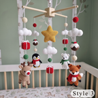 A Christmas-themed baby mobile with crocheted animal and holiday motifs hanging above a crib. The mobile includes a wooden star, white clouds, red and cream beads, gift boxes, a penguin, a bear, a fox, and a reindeer.