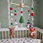 A Christmas-themed baby crib mobile hanging in a nursery. The mobile features crocheted plush animals including a brown reindeer, white polar bear, black penguin, and orange fox, along with a green tree, gift boxes, and hanging beads.