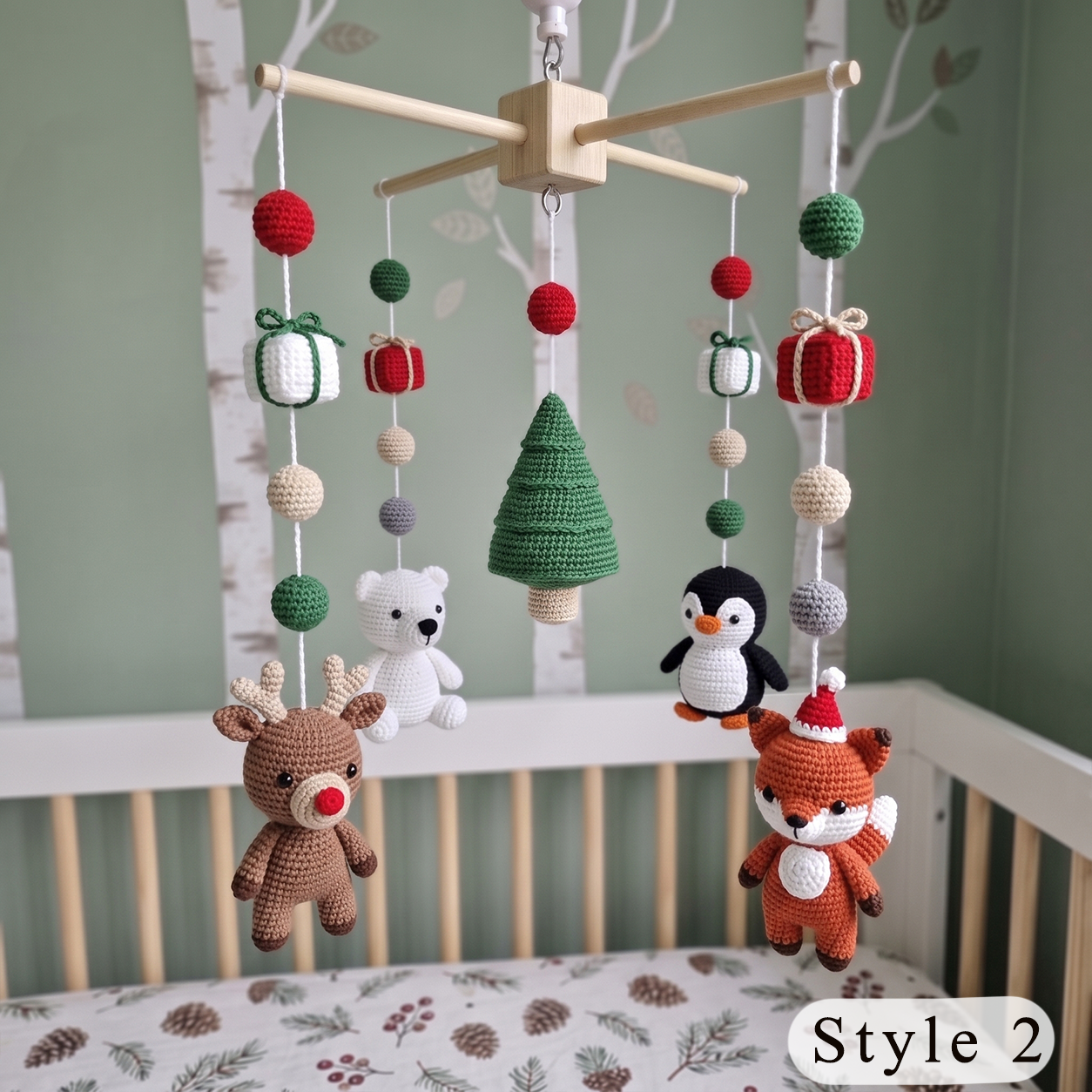 A Christmas-themed baby crib mobile hanging in a nursery. The mobile features crocheted plush animals including a brown reindeer, white polar bear, black penguin, and orange fox, along with a green tree, gift boxes, and hanging beads.