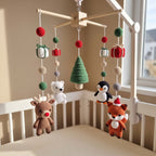 A crocheted baby mobile with Christmas-themed plush toys and ornaments hanging over a white crib in a softly lit nursery. The mobile includes a green tree, red and green beads, gift boxes, a brown reindeer, a white polar bear, a penguin, and an orange fox.