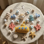 A crocheted baby mobile featuring an ocean theme, including a yellow submarine, octopus, fish, starfish, coral, and seashell charms, displayed on a beige fabric-covered round table.