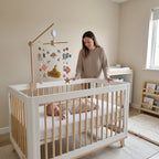 A woman stands beside a white and natural wood baby crib in a softly lit nursery, with a baby lying inside on a white mattress. The crib has a matching mobile with sea-themed hanging decorations, and the room has beige walls, a window, and a small bookshelf.