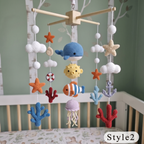 A crocheted baby mobile with an ocean theme, hanging above a crib. The mobile features a wooden crossbar, white yarn clouds, a blue crocheted whale, a yellow sun, an orange and white striped fish, a blue anchor, a purple jellyfish, and various orange starfish and blue coral pieces.