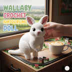 A white crocheted kangaroo amigurumi doll with pink inner ears is shown on a miniature train track next to a cup of latte, with a human hand gently touching its head. Large text in the upper left reads 'WALLABY CROCHET AMIGURUMI DOLL'.