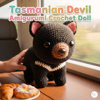 A crocheted Tasmanian Devil amigurumi doll with a black body, pink inner ears, and a cream collar sits on a wooden table by a window. A hand is holding the left side of the doll, and croissants are visible on a plate in the foreground.