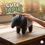 A handmade crocheted tapir doll with a black and white body and pink inner ears, being held by a person's hand on a wooden table with potted plants in the background. Text overlay reads 'CUTE TAPIR HANDMADE DOLL'.