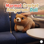 A crocheted marmot doll sits on a blue train seat with a human hand resting on its head. Large stylized text at the top reads 'Marmot Crochet Amigurumi Doll'.