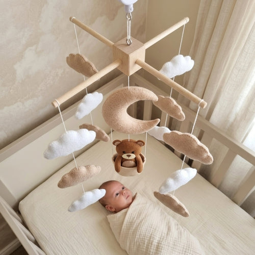 A baby mobile with a wooden star frame, hanging felt clouds, a crescent moon, and a teddy bear, shown above a baby lying in a crib.