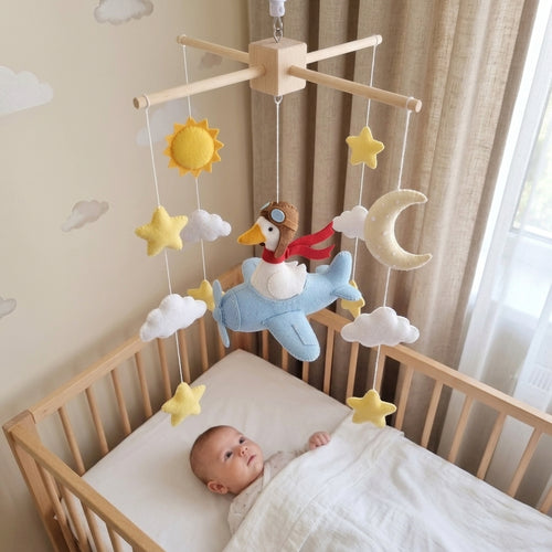 A baby mobile with a wooden cross frame hangs over a baby crib. The mobile features a light blue airplane with a toy bird wearing aviator goggles and a red scarf, surrounded by yellow stars, a yellow sun, white clouds, and a cream-colored crescent moon.