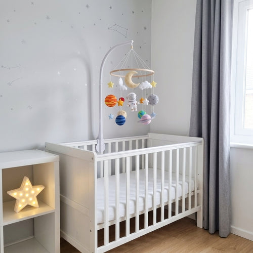 A white wooden baby crib in a softly lit nursery, with a white arched mobile above it holding colorful planets, stars, and a moon. A matching white bedside shelf with a glowing star lamp sits beside the crib, and gray curtains hang by a window.