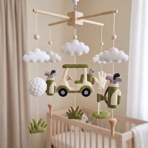 A baby nursery mobile featuring felt golf-themed decorations including a golf cart, golf bags, clubs, clouds, grass, and a golf ball, hanging above a light wood crib in a softly lit room.