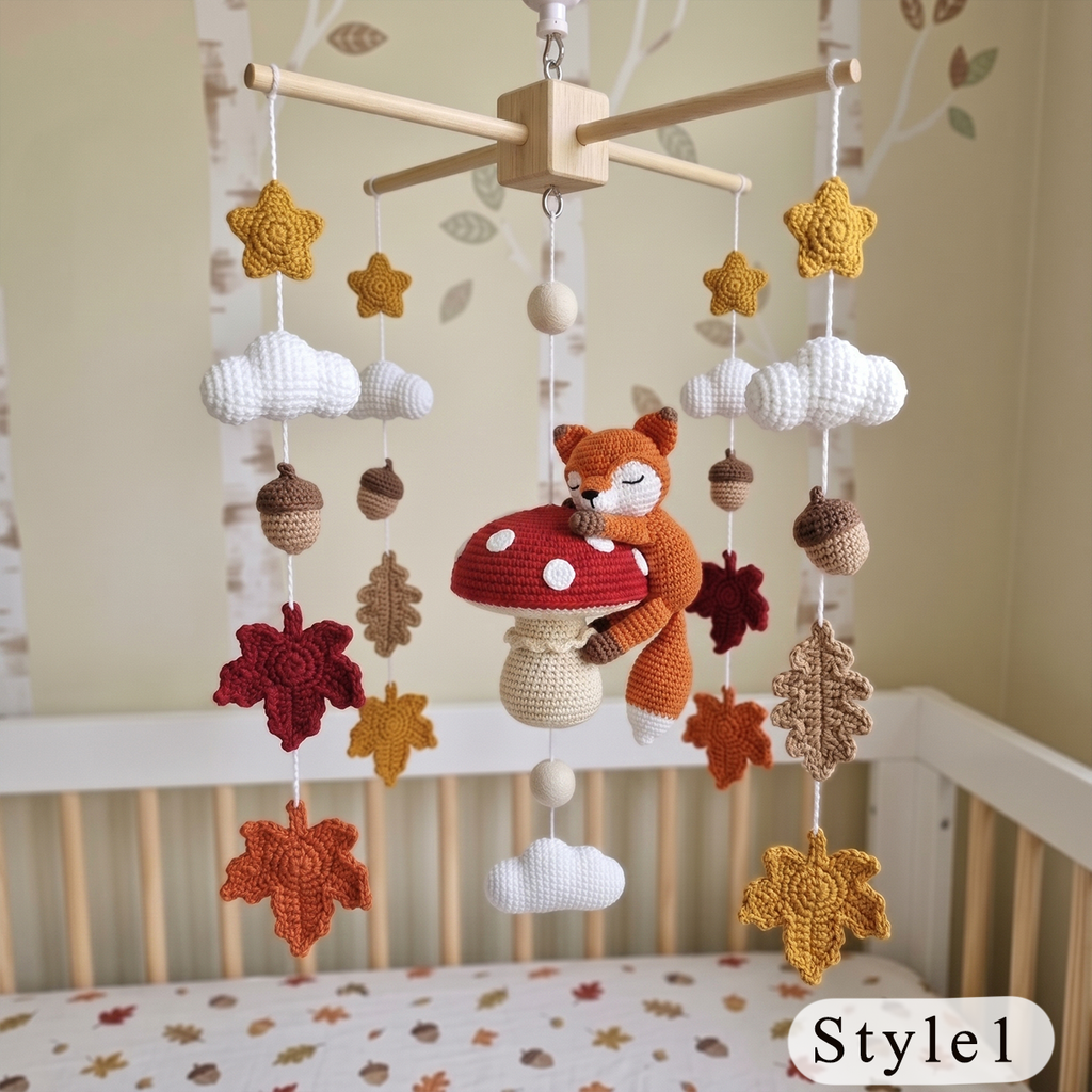 A crocheted baby mobile with a fox, mushroom, clouds, stars, acorns, and autumn leaves hanging above a crib in a nursery. The mobile has a natural wood top frame and white, beige, orange, red, brown, and yellow details.