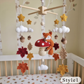 A crocheted baby mobile with a fox, mushroom, clouds, stars, acorns, and autumn leaves hanging above a crib in a nursery. The mobile has a natural wood top frame and white, beige, orange, red, brown, and yellow details.