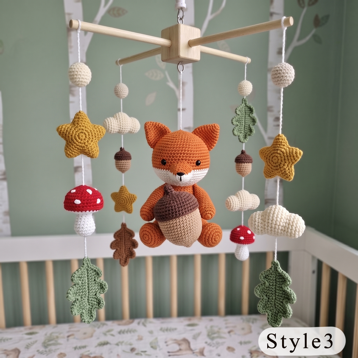 A crocheted baby mobile hanging above a crib, featuring an orange and beige plush fox holding an acorn, surrounded by hanging stars, leaves, acorns, clouds, and red-and-white mushrooms. The crib is set against a soft green wall with birch tree decals, and a white 'Style3' label appears in the bottom right corner.