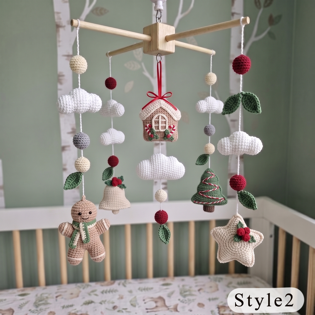 A wooden baby crib with a hanging crocheted mobile featuring a gingerbread man, Christmas tree, house, clouds, bells, and red pom-poms, shown in a nursery setting with a green wall and birch tree print bedding.