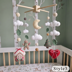 A wooden baby crib with a crocheted hanging mobile featuring clouds, a crescent moon, leaves, a gingerbread man, a house, and a red heart ornament, set against a soft green wall.