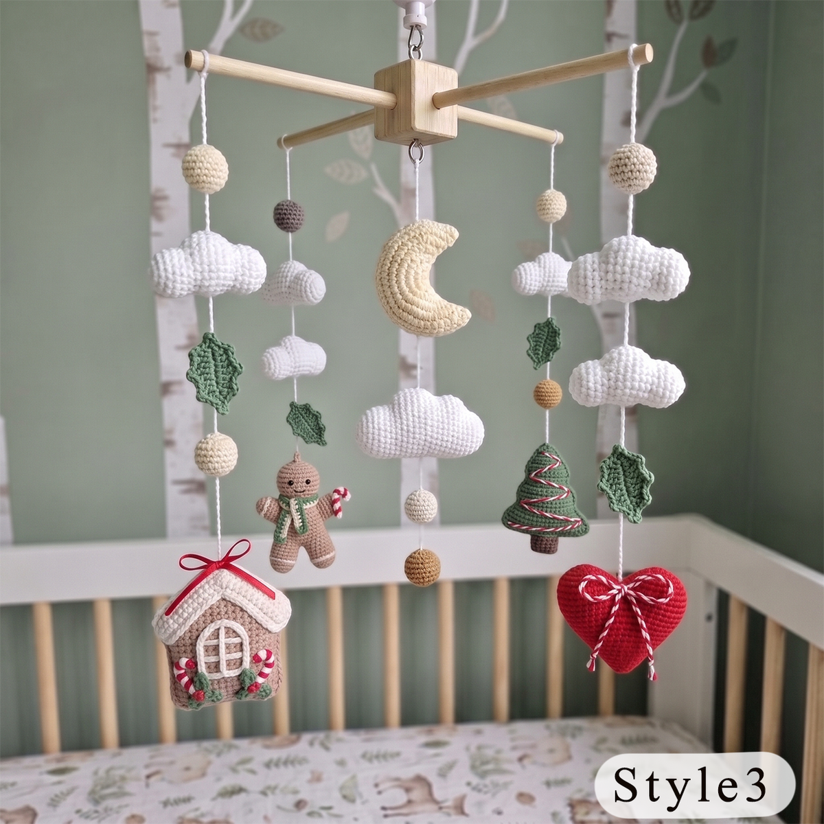 A wooden baby crib with a crocheted hanging mobile featuring clouds, a crescent moon, leaves, a gingerbread man, a house, and a red heart ornament, set against a soft green wall.