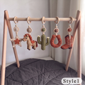 A wooden baby gym with five crocheted hanging toys shaped like a star, horse, cactus, U-shaped ring, and cowboy boots, displayed on a dark gray quilted blanket against a light gray background.