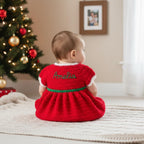 A baby sits on a cream knit rug in a living room, viewed from behind wearing a red crocheted dress with white trim, a green waistband, and the name 'Amelia' embroidered in green on the back. A decorated Christmas tree is visible on the left.