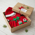 A handmade Christmas-themed crochet pet dress and matching hat presented in an open kraft gift box with the lid leaning behind it. The dress is bright red with a green bow, white trim, and small white paw-print embellishments, while the hat has a white trim.