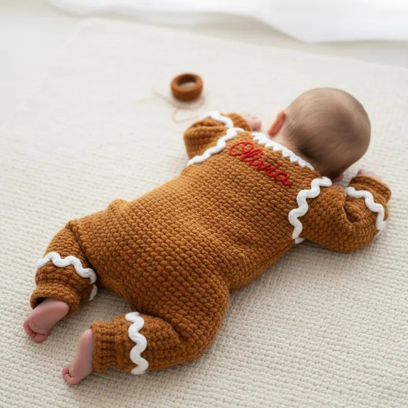 A baby lying on their tummy on a white textured blanket, seen from behind, wearing a handmade brown crochet gingerbread romper with white icing-style trim and the custom name Olivia embroidered in red across the upper back.