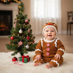 A cute smiling baby wearing a handmade brown crochet gingerbread romper and matching hat, sitting on a fluffy white rug beside a decorated mini Christmas tree and holiday gifts with a cozy fireplace background.