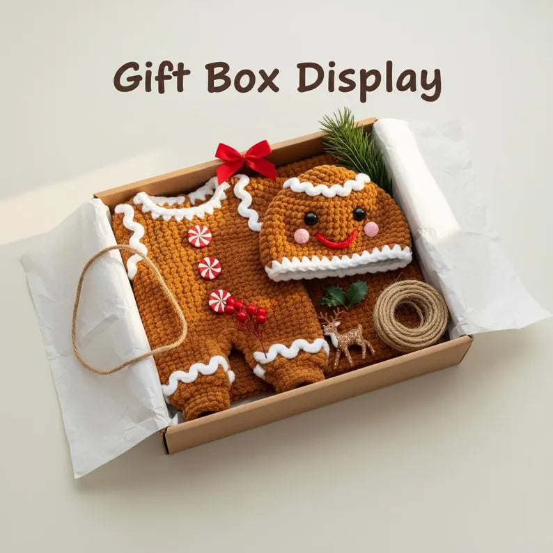 A handmade brown crochet gingerbread baby romper and matching smiling hat neatly arranged inside an open craft gift box with red bow, pine sprig, twine roll, and miniature deer decor, with overlay text reading Gift Box Display.