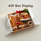 A handmade brown crochet gingerbread baby romper and matching smiling hat neatly arranged inside an open craft gift box with red bow, pine sprig, twine roll, and miniature deer decor, with overlay text reading Gift Box Display.