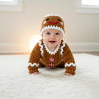 A cute baby crawling on a soft white rug wearing a handmade brown crochet gingerbread romper with white icing trim and peppermint buttons, paired with a matching gingerbread face hat.