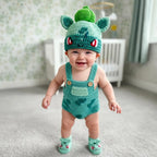 A smiling baby wearing a handmade crochet outfit inspired by Bulbasaur, featuring a green hat with bulb details, teal-green overalls with brown buttons, and matching booties, standing in a softly lit nursery.