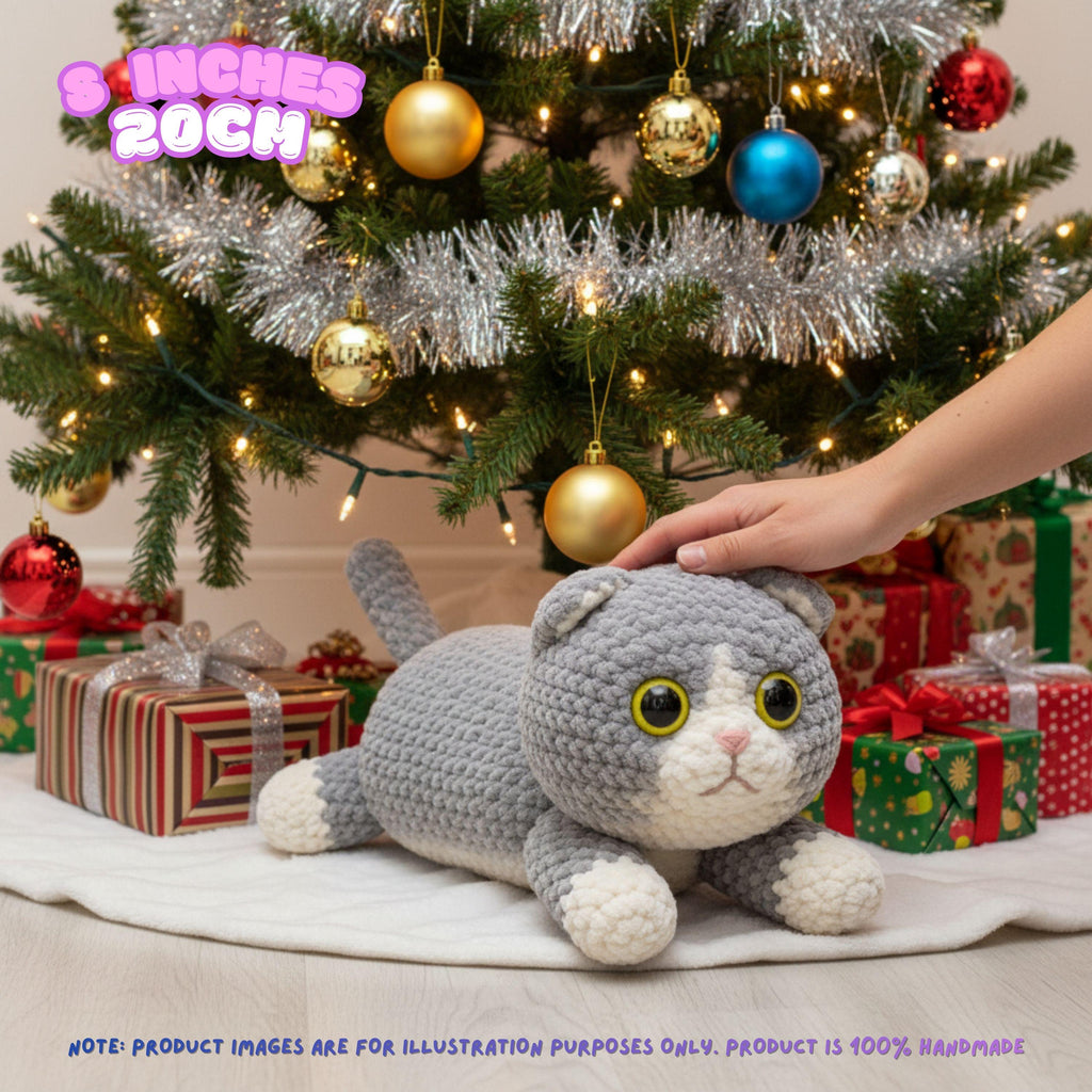A gray and white crocheted cat plush toy lying on a white blanket under a decorated Christmas tree, with a human hand touching its head and wrapped gifts around its base. Text overlays indicate the size as 8 inches / 20 cm.