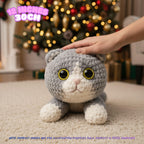 A gray and white crocheted cat plush toy with large yellow eyes sits on a beige carpet in front of a blurred Christmas tree. A human hand is resting on the cat's head, and text in the top left reads '12 INCHES 30CM'.