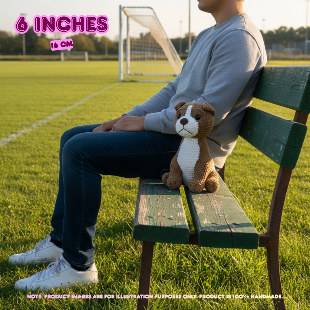 A person sitting on a green wooden bench on a grassy soccer field, holding a small brown and white crocheted dog toy. Text overlays indicate the toy is 6 inches (15 cm) and note that the product image is for illustration purposes only.