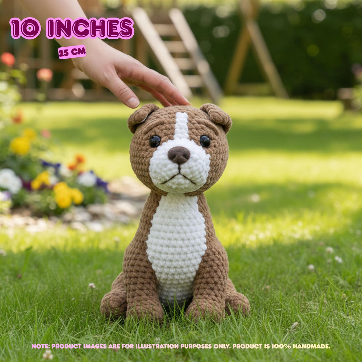 A crocheted dog toy sits on green grass in a garden, with a human hand reaching toward its head. The toy is brown and white with black eyes and a brown nose.