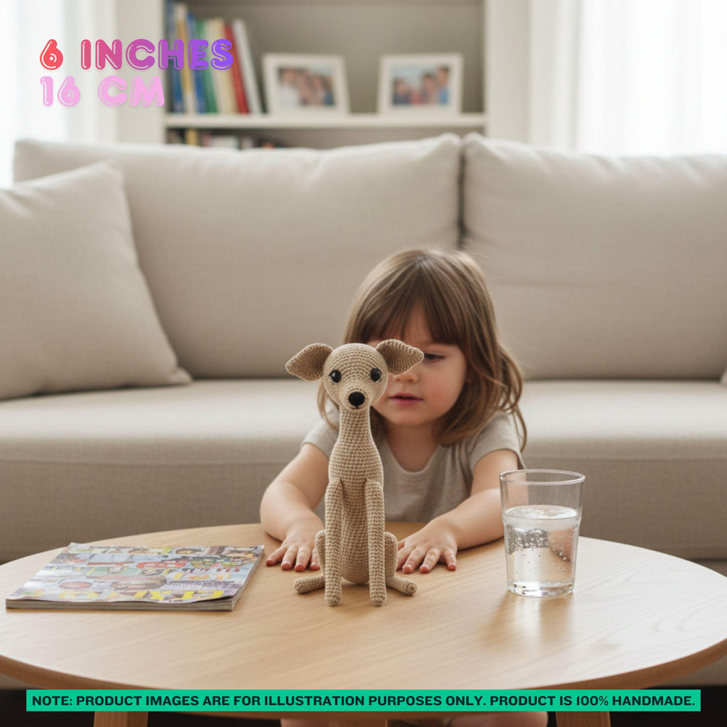 A young child sits at a round wooden table in a living room setting, looking at a small crocheted dog toy. The child is wearing a light gray t-shirt and there is a glass of water and a magazine on the table.