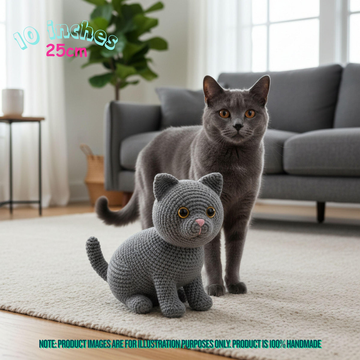 A gray real cat stands behind a crocheted plush cat toy in a bright living room. The image includes size text reading 10 inches / 25 cm and a note at the bottom stating the product image is for illustration purposes only and the product is 100% handmade.