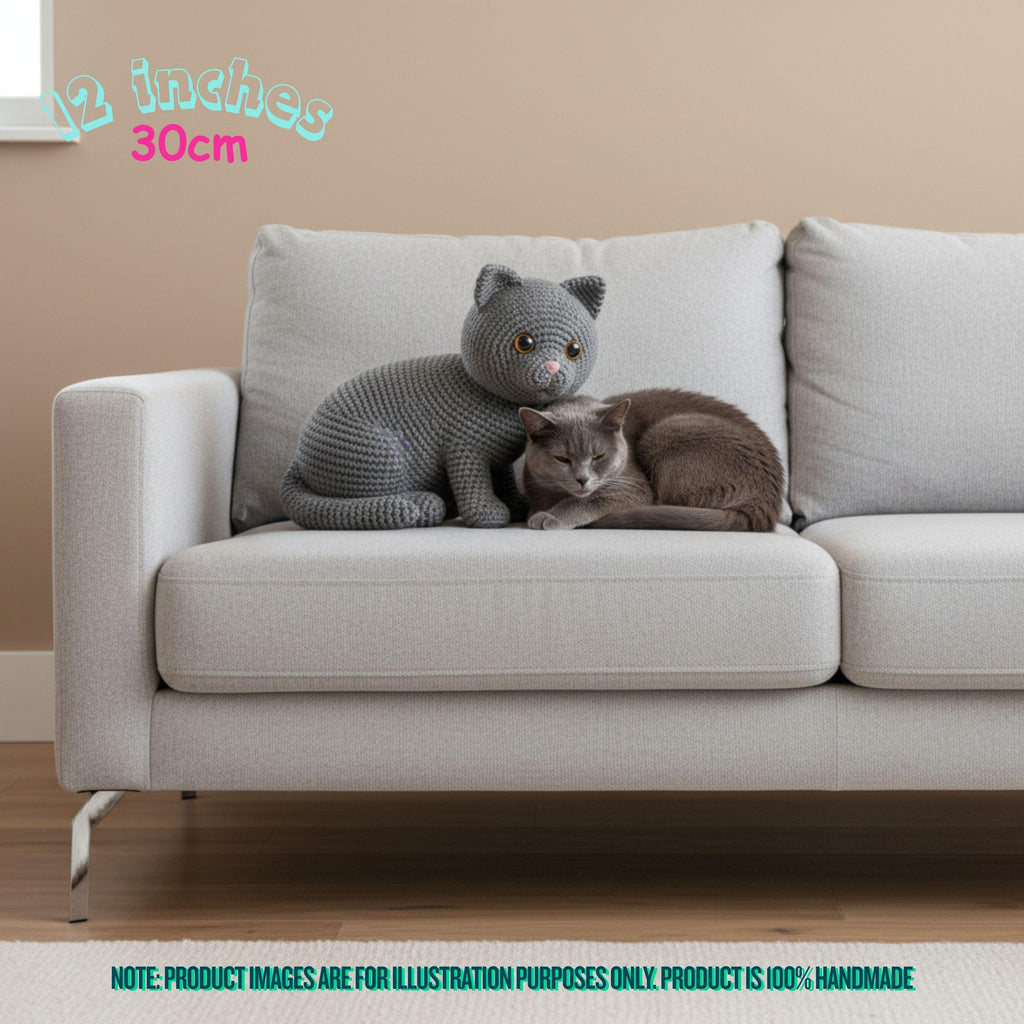 A crocheted cat-shaped cushion or pillow displayed on a light gray sofa, with a real cat resting beside it. The photo is taken in a living-room setting with a beige wall and wood floor, and text in the image notes 12 inches / 30 cm and a note that product images are for illustration purposes only.