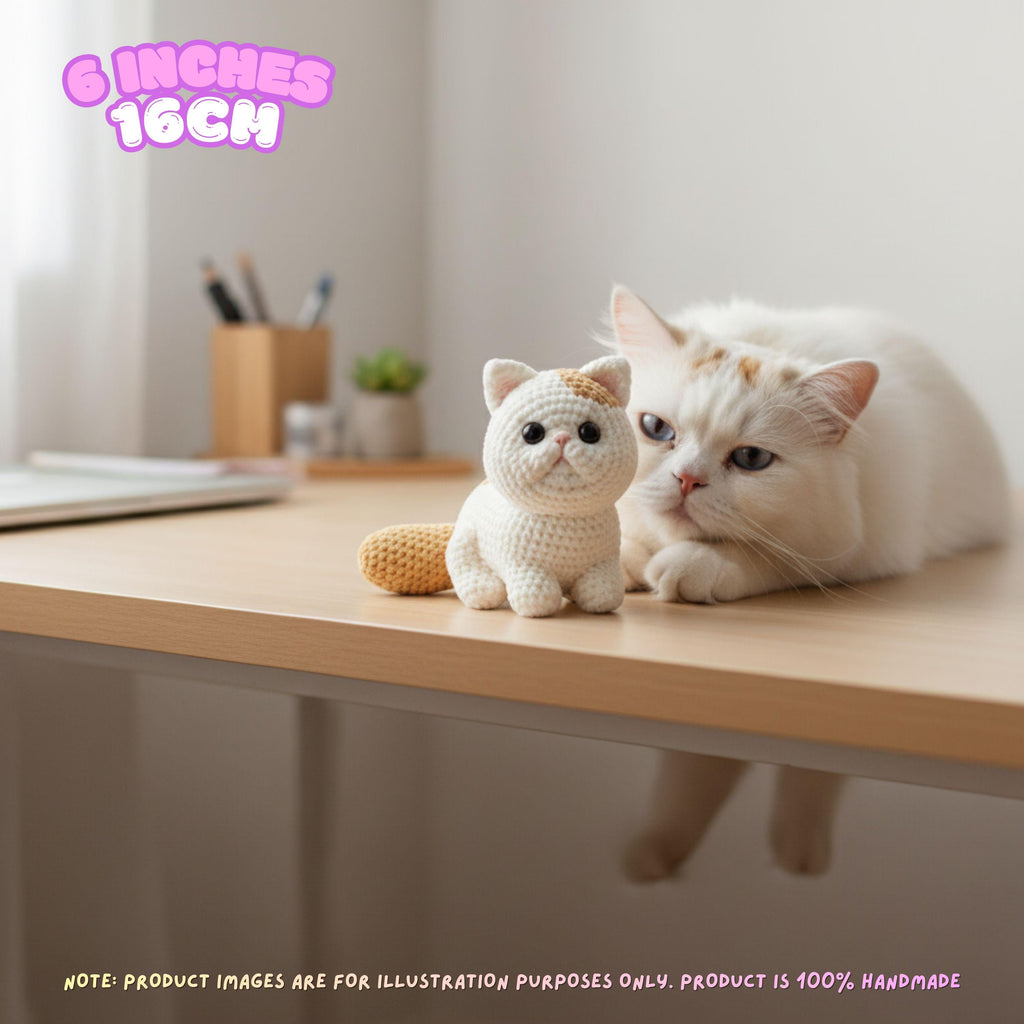 A white cat lies on a light wood desk next to a small crocheted cat toy. The scene is indoors with soft natural light, and a pink text overlay at the top left reads '6 INCHES 16CM'.